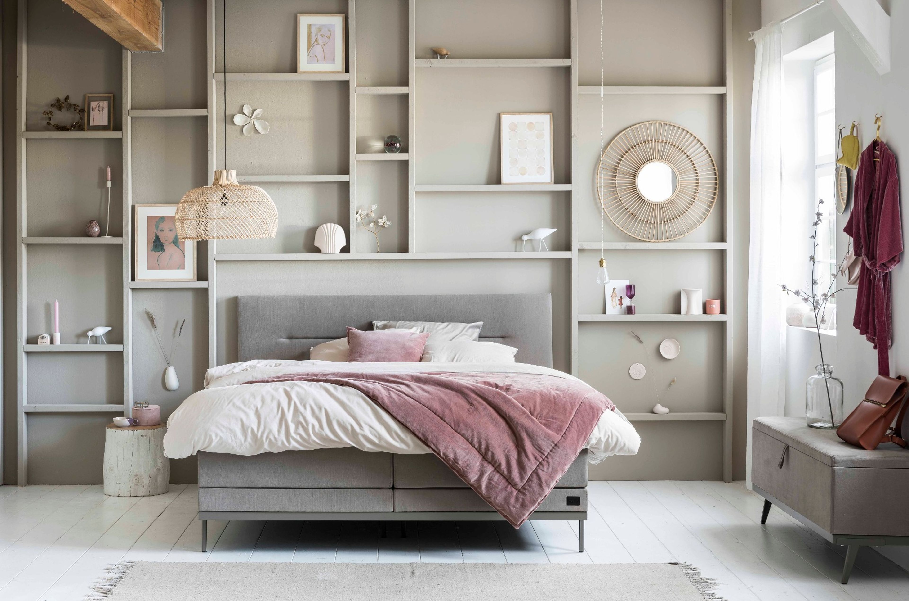 Helles Boho Schlafzimmer mit grauem Boxspringbett, rosa Akzenten, Rattan-Dekoration und offenen Regalen im natürlichen Wohnstil.